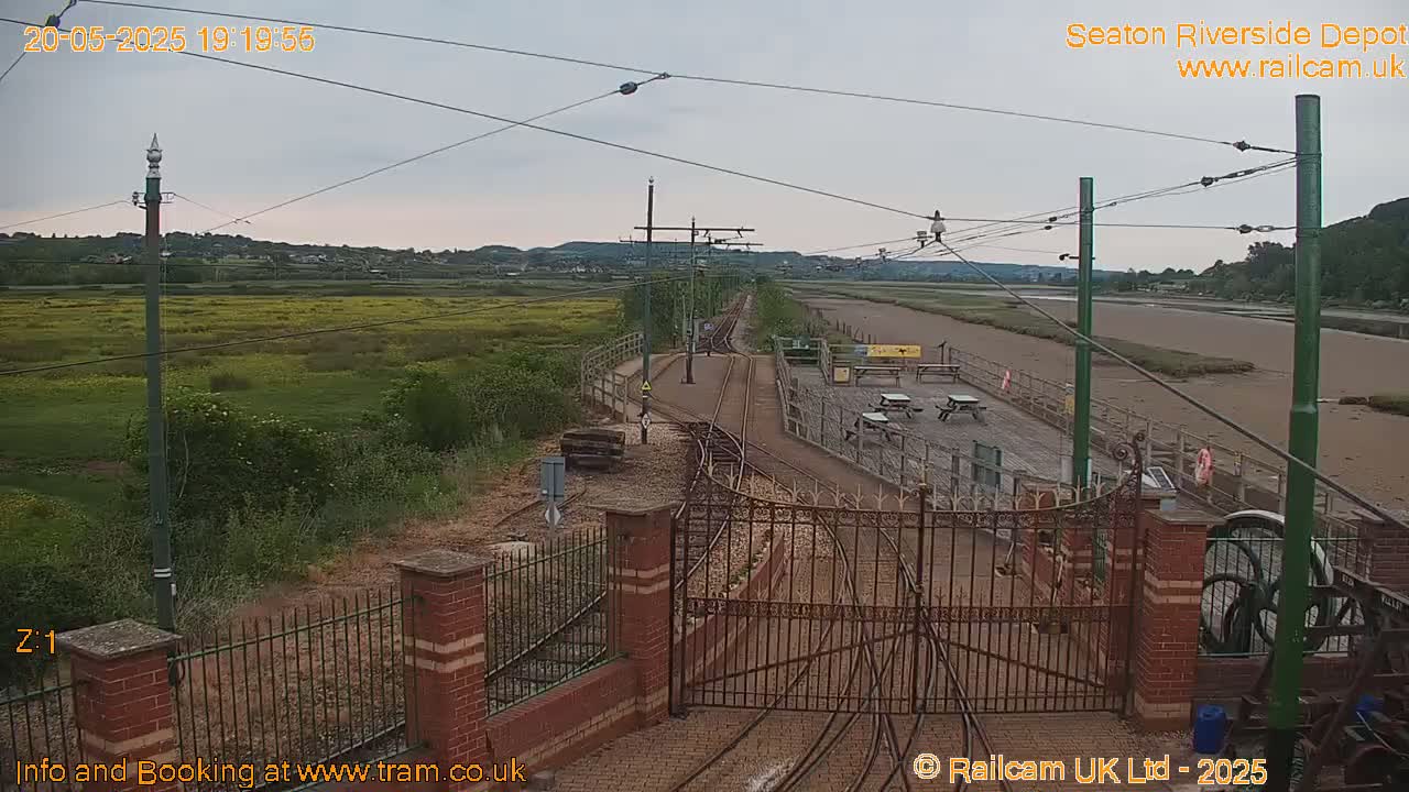 A train depot with tracks, a fenced area, and a muddy riverbank under an overcast sky.