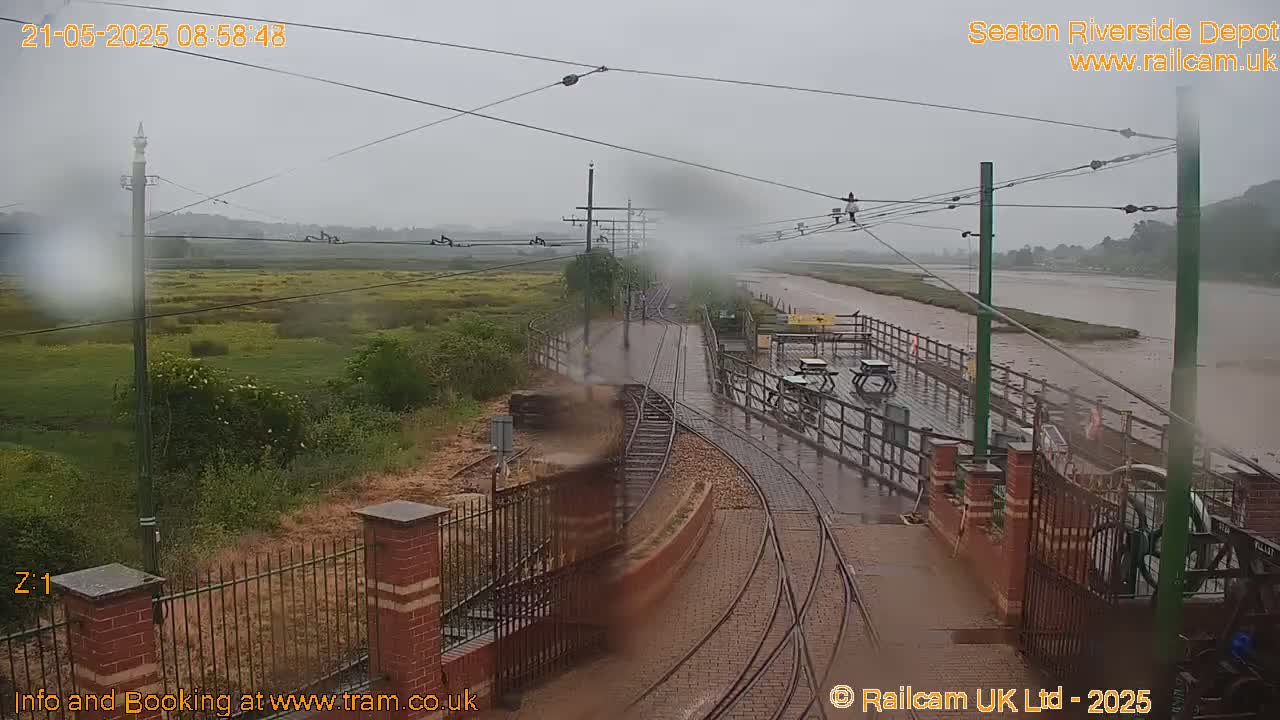 A rain-soaked railway depot with tram tracks and a platform overlooks a muddy river under a gray sky.