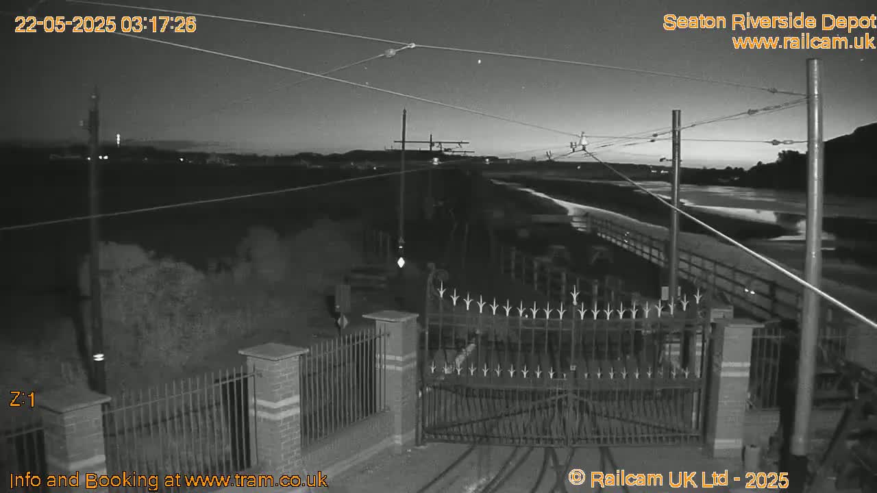 A nighttime, low-light grayscale image shows an empty train depot with a closed gate, power lines, and a body of water visible in the background.