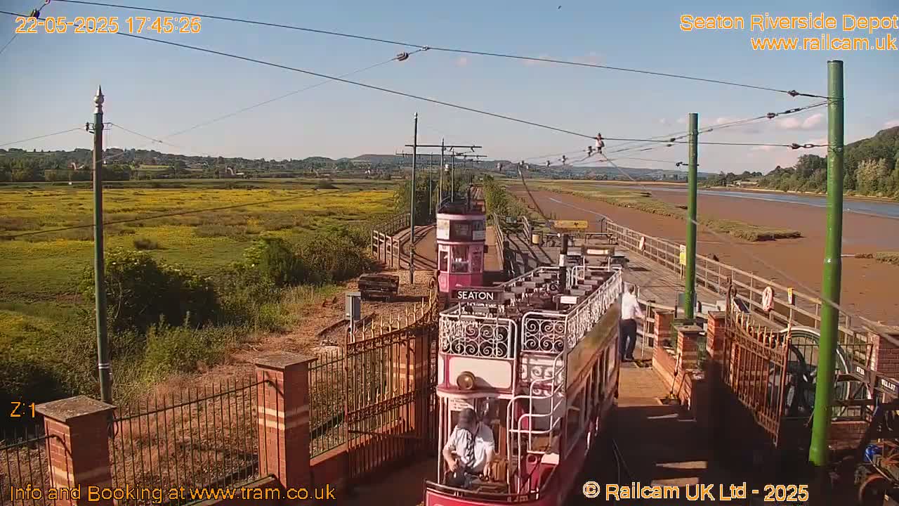 Two vintage trams are on a track next to a river under a clear, sunny sky; a person stands near one of the trams and yellow wildflowers are visible in the distance.