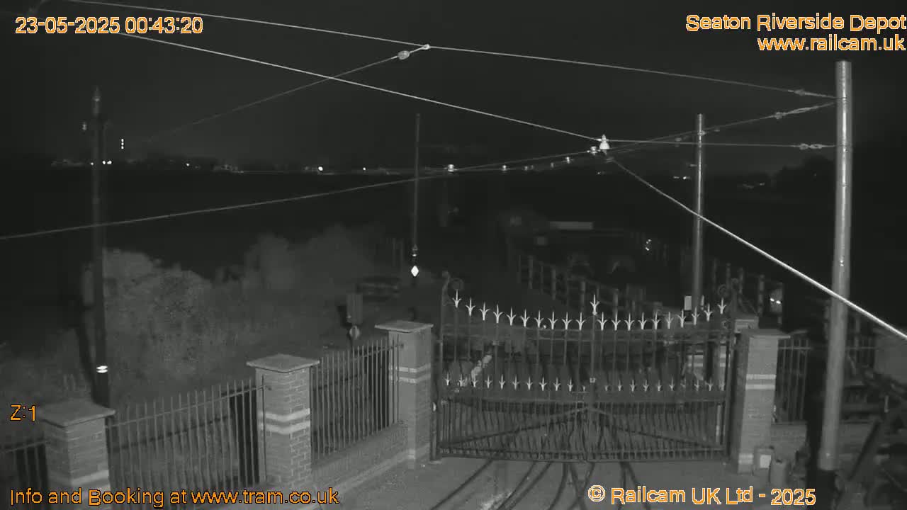 A nighttime, black-and-white view of a closed gate at a depot, with overhead wires and some vegetation visible.