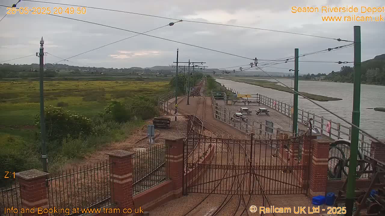A railway depot with a gated entrance overlooks a calm river under an overcast sky, with a grassy area and wildflowers visible in the background.