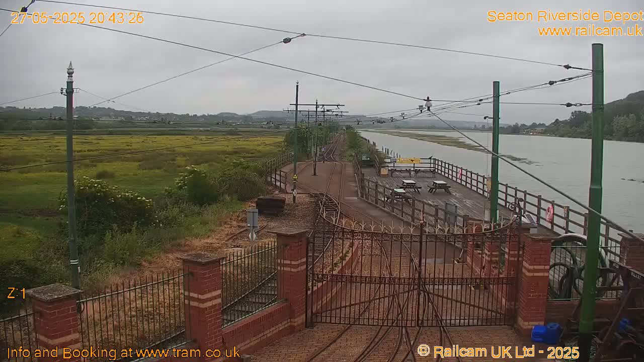 A view of a railway line next to a river on an overcast day, with a gated entrance and a wooden platform with picnic tables visible.