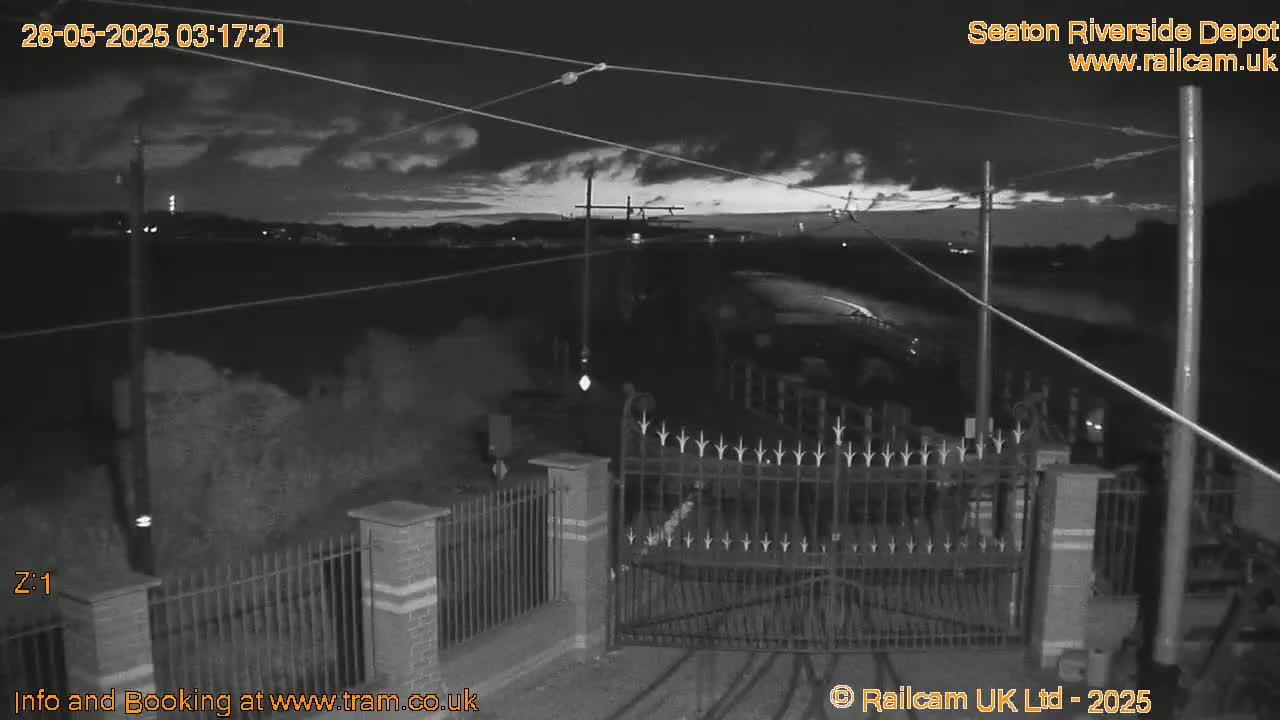 A nighttime, overcast scene shows a closed gate and fence in front of a railway depot with overhead power lines, and a dimly lit road in the background.