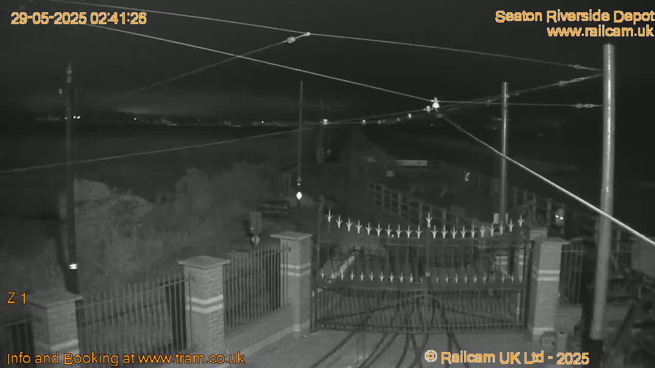 A nighttime, black-and-white view of a gated area with overhead wires, possibly a depot, under a dark sky.