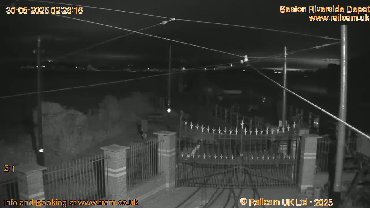 A nighttime, low-light view of a gated entrance to what appears to be a depot, with overhead wires and a distant, faintly illuminated horizon.