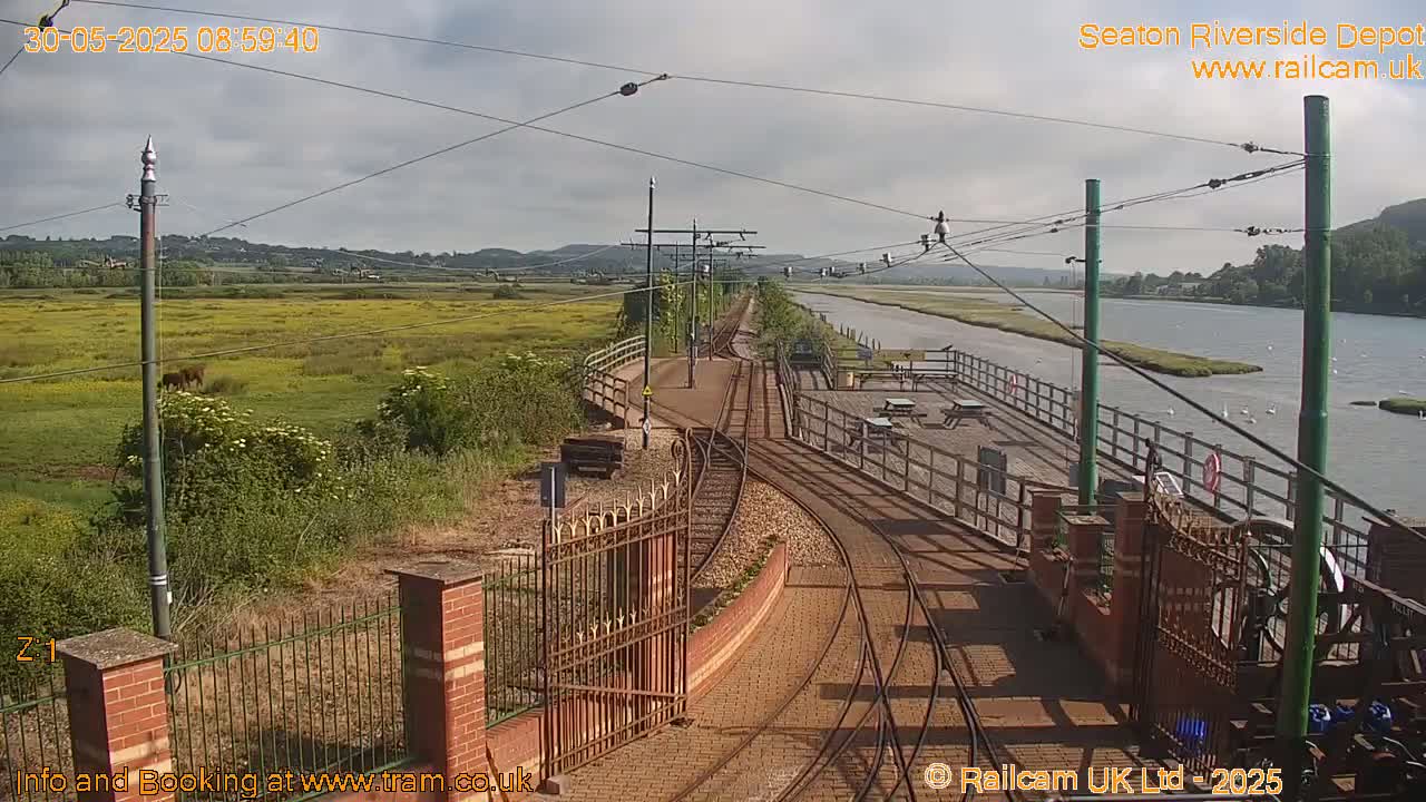 A railway track curves alongside a river under a partly cloudy sky, with a grassy area and a fence visible in the foreground.