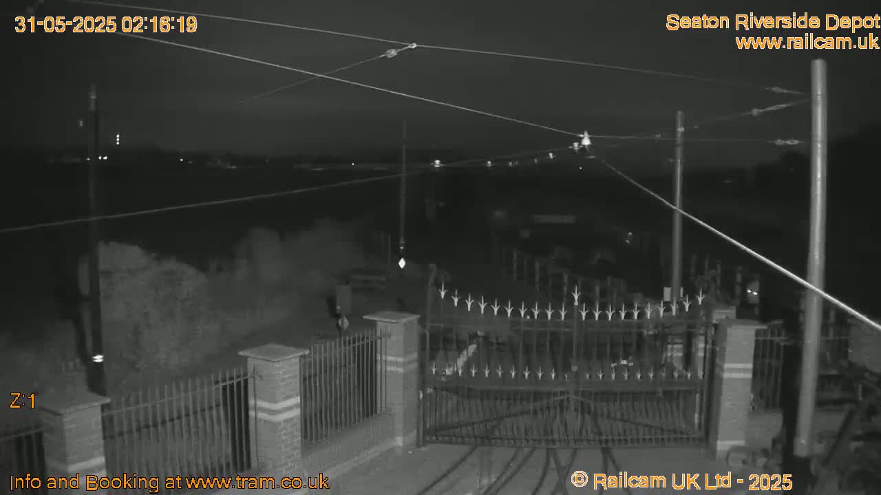 A nighttime, low-light view of a gated entrance to a depot, with overhead wires and a mostly dark surrounding area.