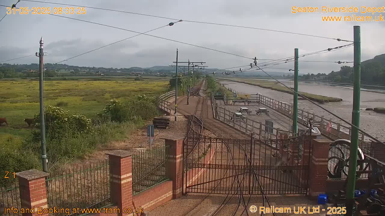 A train depot with railway tracks and a gate, situated beside a river under an overcast sky, and cows are visible in a field beyond.