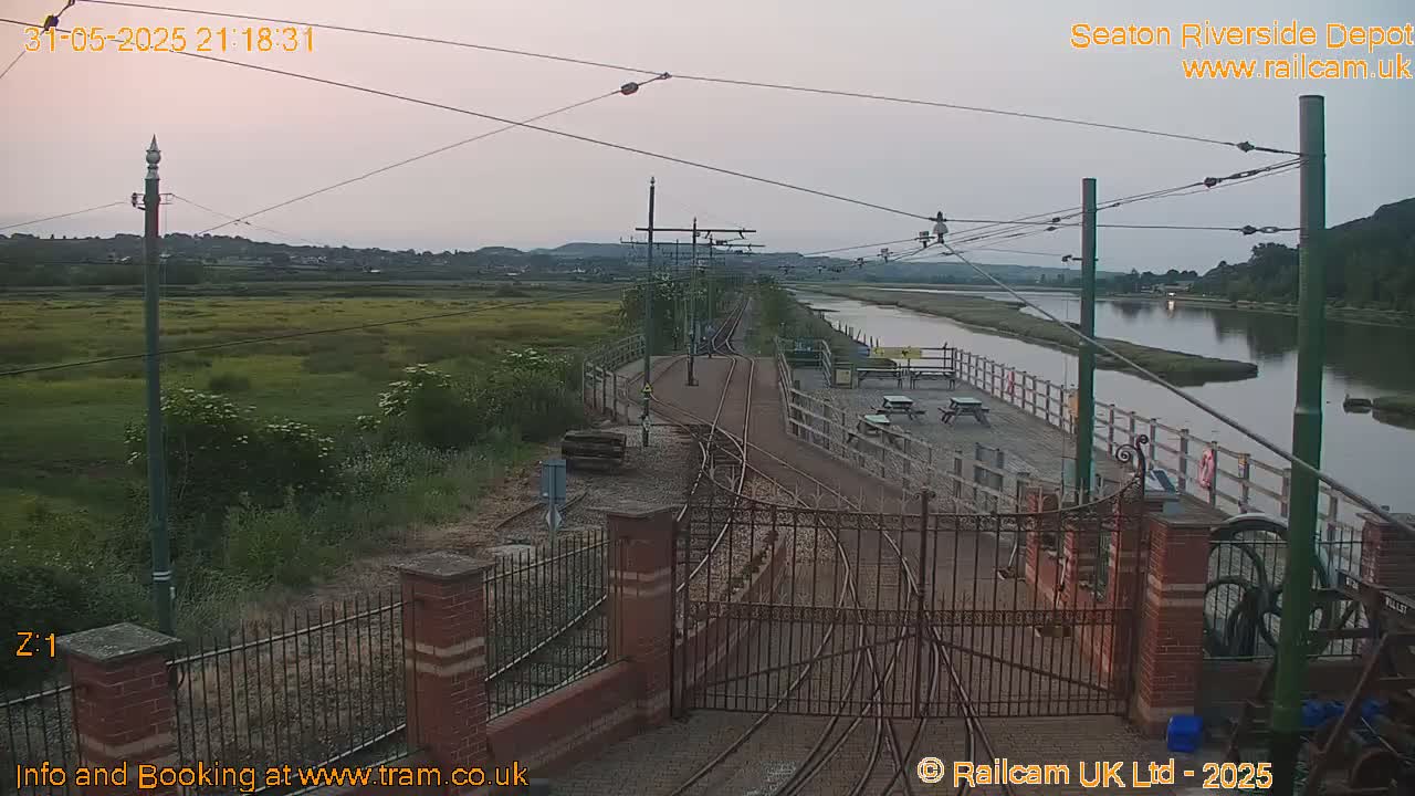 A train depot with railway tracks and gates is situated by a calm river under a dusky sky.