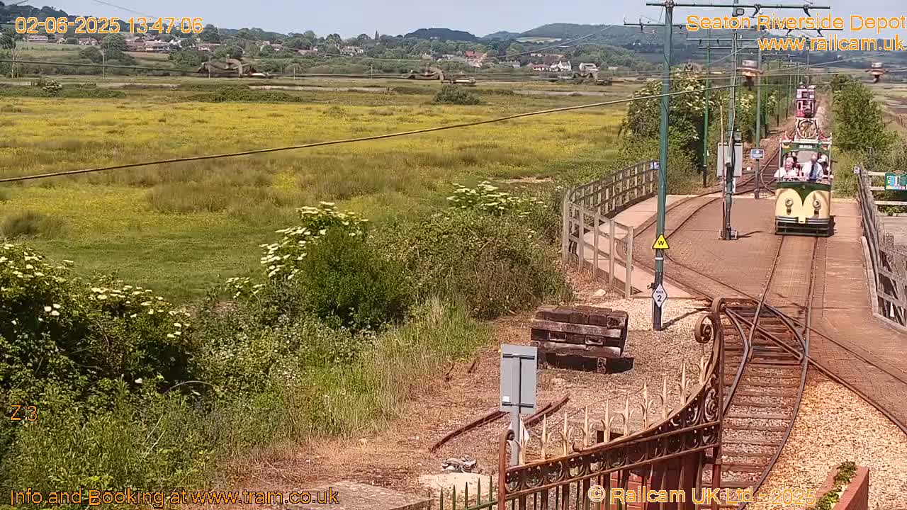 A passenger tram travels along a railway track in a sunny, grassy field, with a village visible in the distance.