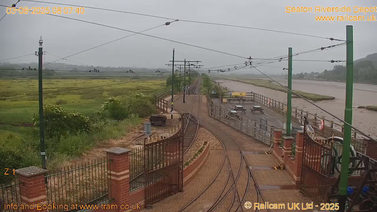 A railway depot with a tram line and overhead wires is seen on an overcast day next to a wide, muddy river and a marshy area.