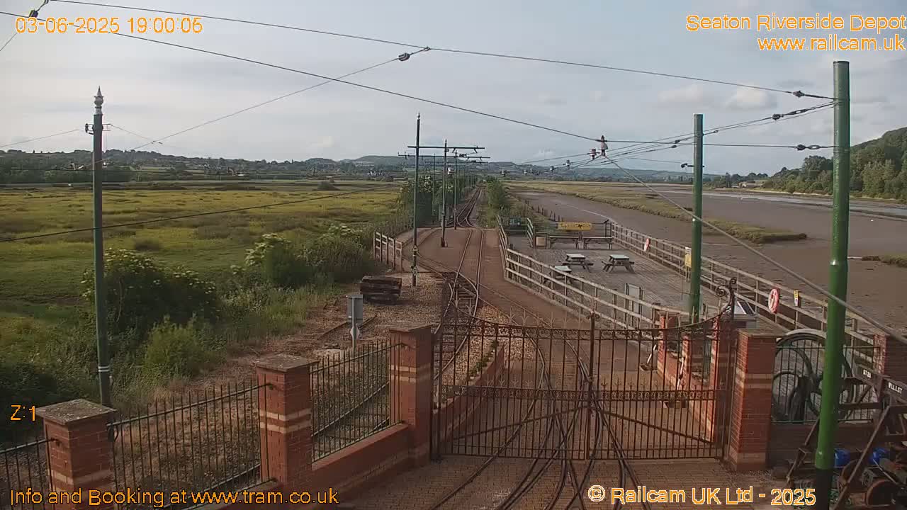 A train depot with railway tracks and a gate, overlooking a flat expanse of land and a waterway under a partly cloudy sky.