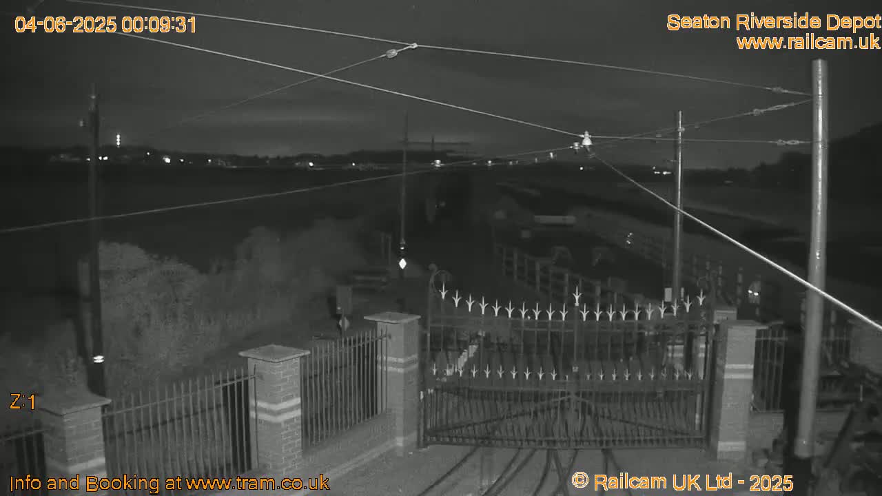A nighttime, low-light view shows an open metal gate at a depot, with overhead wires and a brick fence visible under a dark sky.