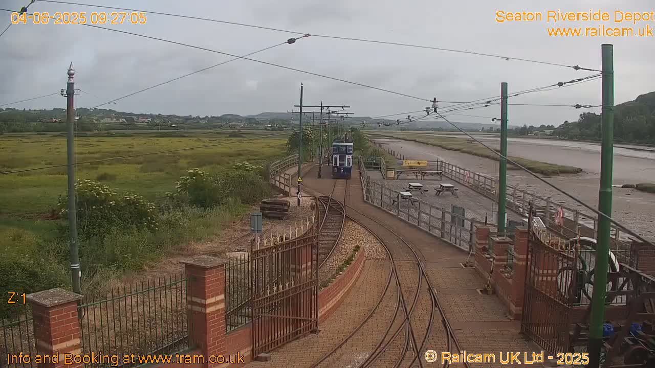A small tram is approaching a station on a cloudy day, beside a river with exposed mudflats.