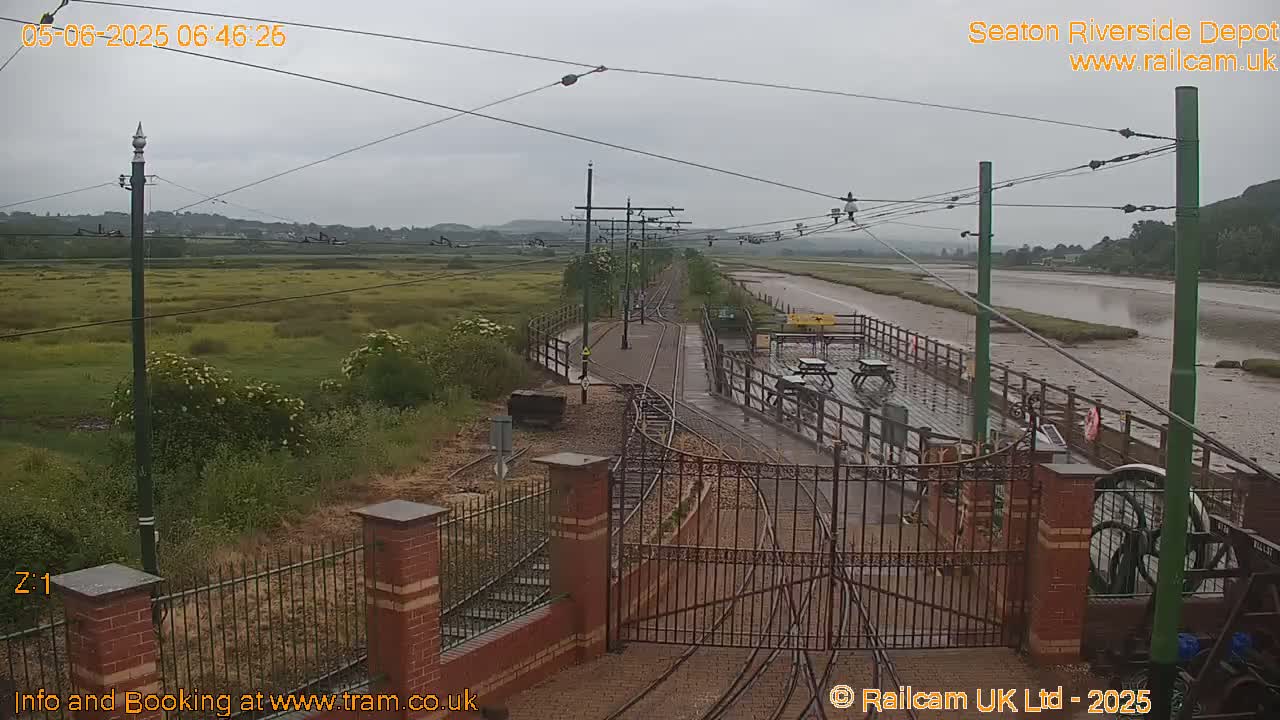 A view of a train depot on an overcast day, showing train tracks, a gate, and a muddy river in the background.