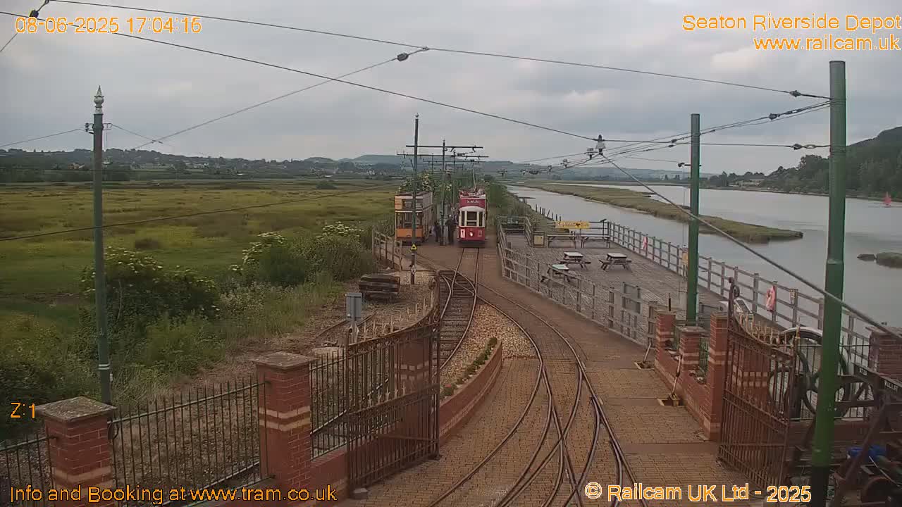 Two vintage trams are parked at a depot near a river under an overcast sky.