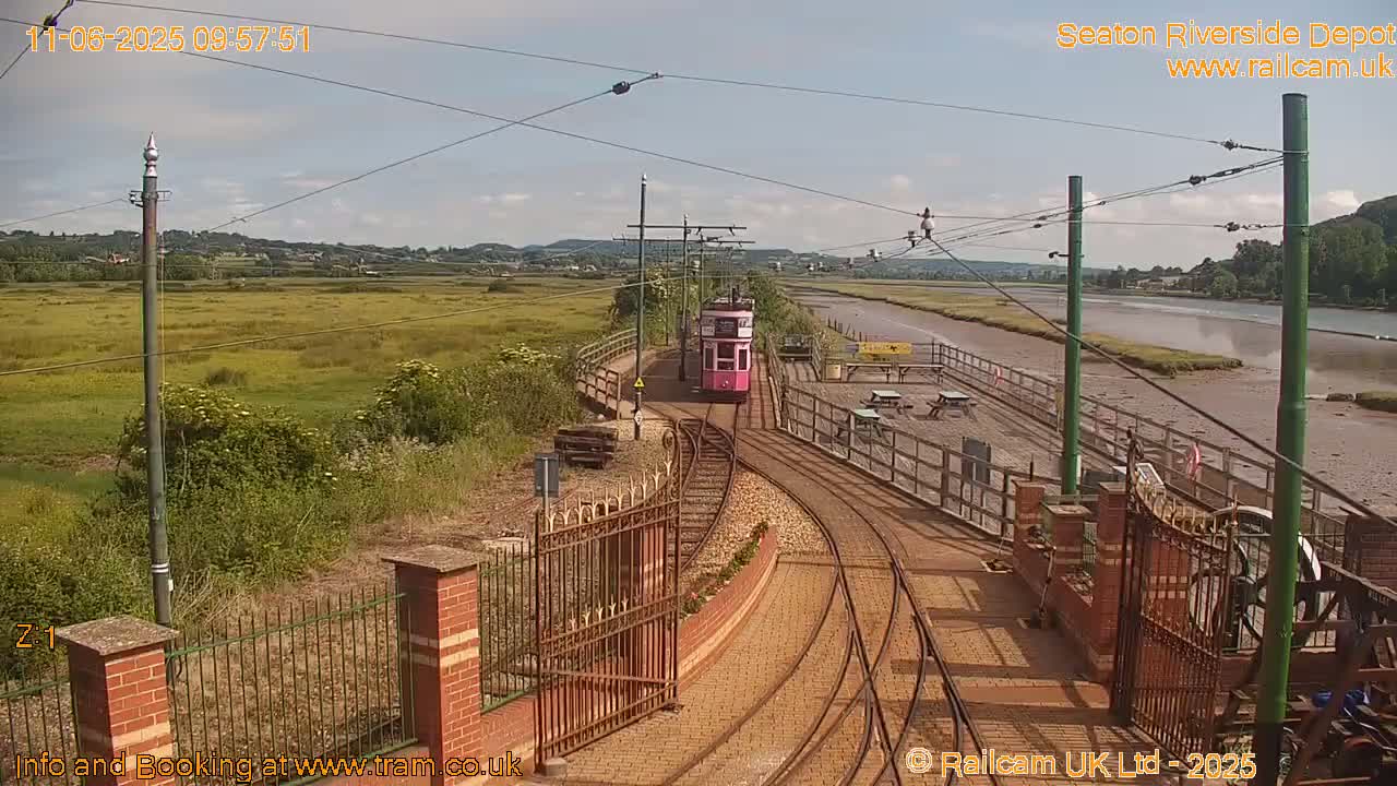 A pink tram travels on tracks toward a depot by a tidal river under a mostly sunny sky.
