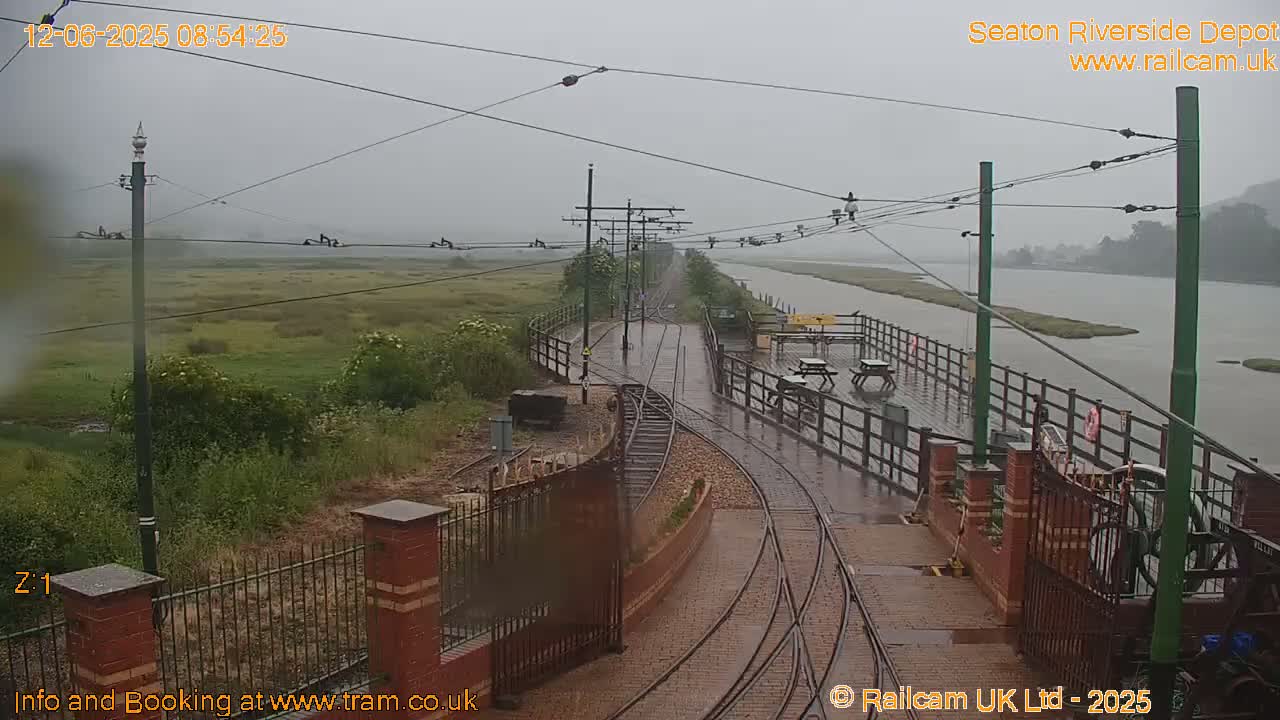 A rain-soaked tram depot platform with railway tracks curves toward a body of water under an overcast sky.