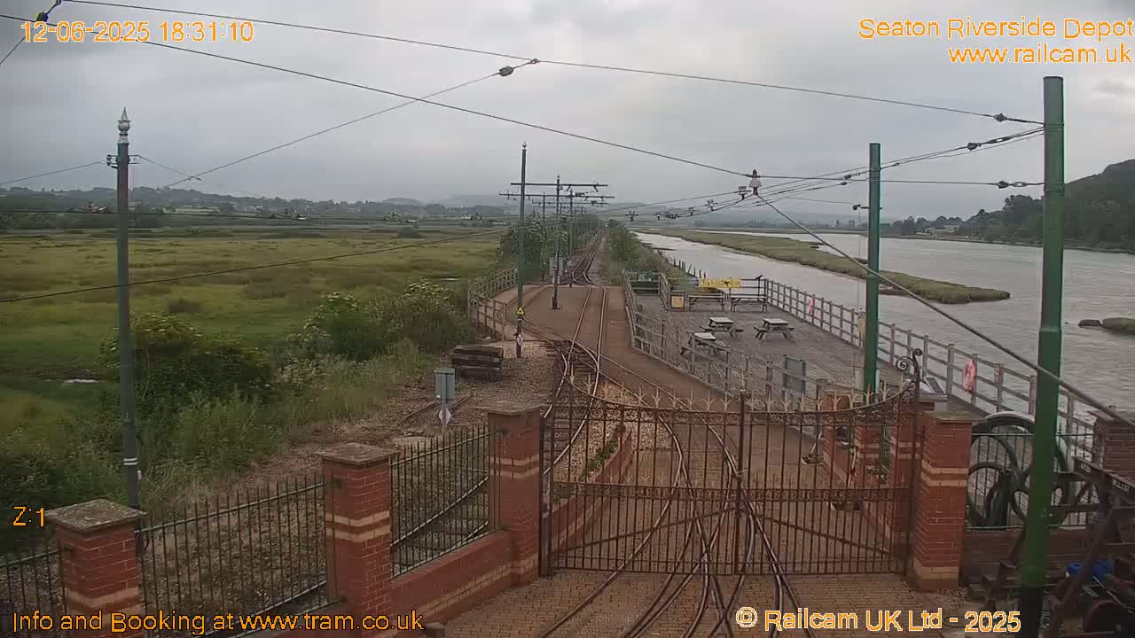 A railway track and depot are seen next to a calm river under an overcast sky.