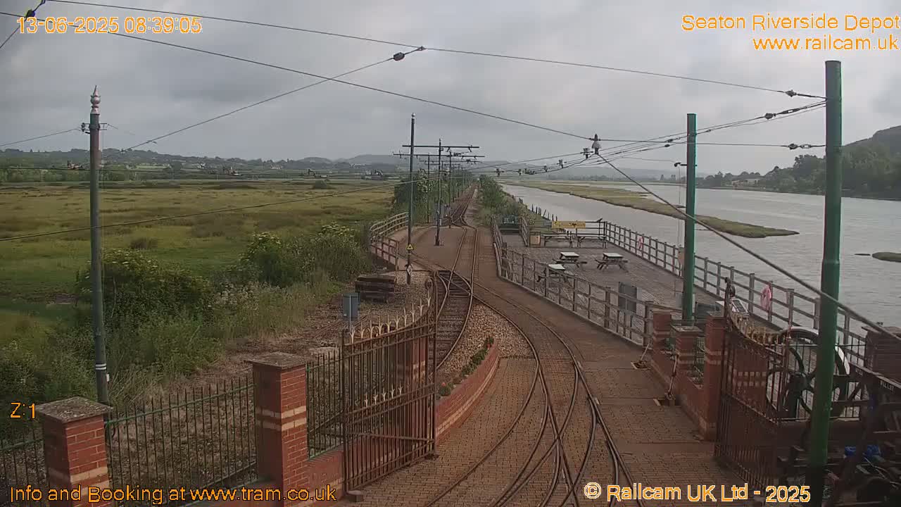 Train tracks curve alongside a river under an overcast sky, leading to a depot with a wooden platform and picnic tables.