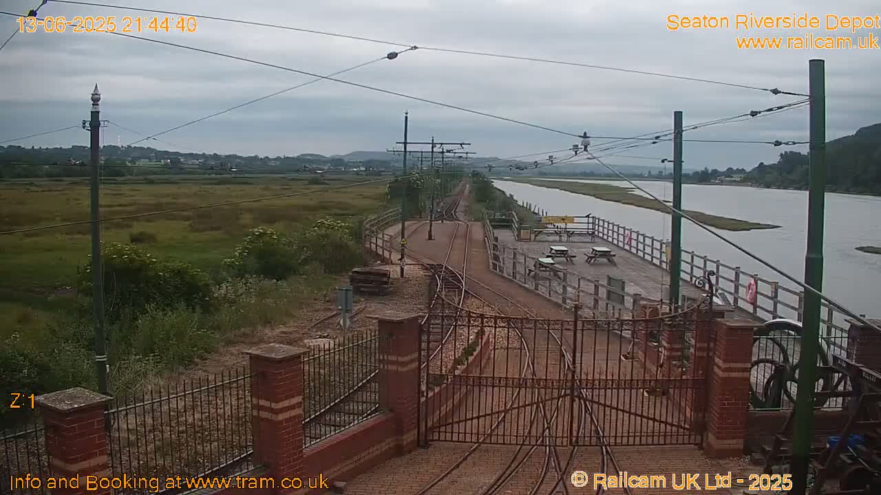 A railway depot with tracks and gates is situated next to a calm river under an overcast sky.