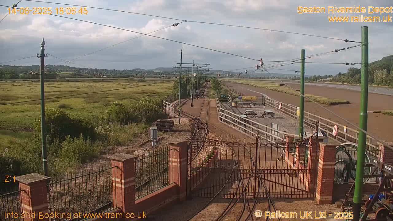 A train depot with tracks and a gated entrance is situated beside a muddy, low-tide estuary under a partly cloudy sky.