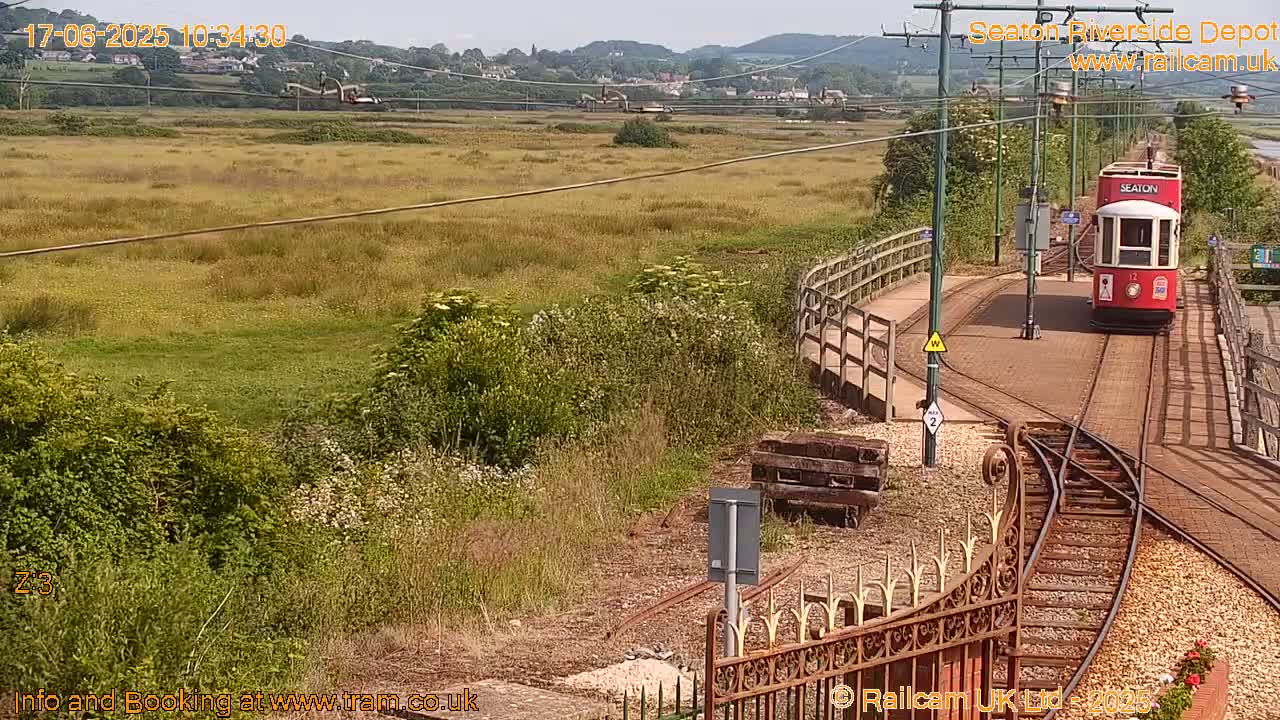A red tram approaches a railway junction in a grassy area on a sunny day, with hills visible in the distance.