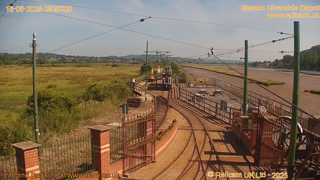 A sunny day at a tram depot shows a tram on the tracks with a flat expanse of tidal mudflats and grassy areas in the background.