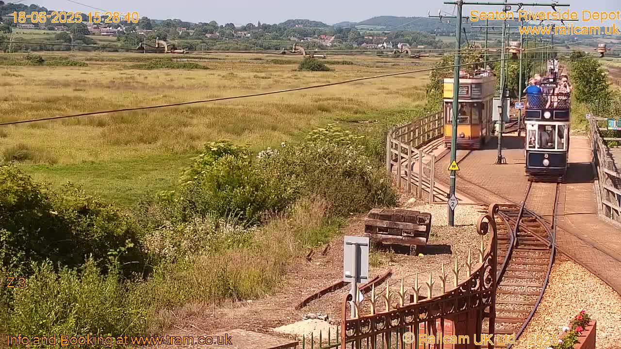 Two vintage trams are traveling along a railway track in a sunny, grassy field, with a distant village visible in the background.