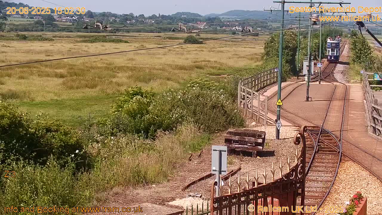 A tram approaches a railway junction on a sunny day, with a grassy field and distant hills visible in the background.