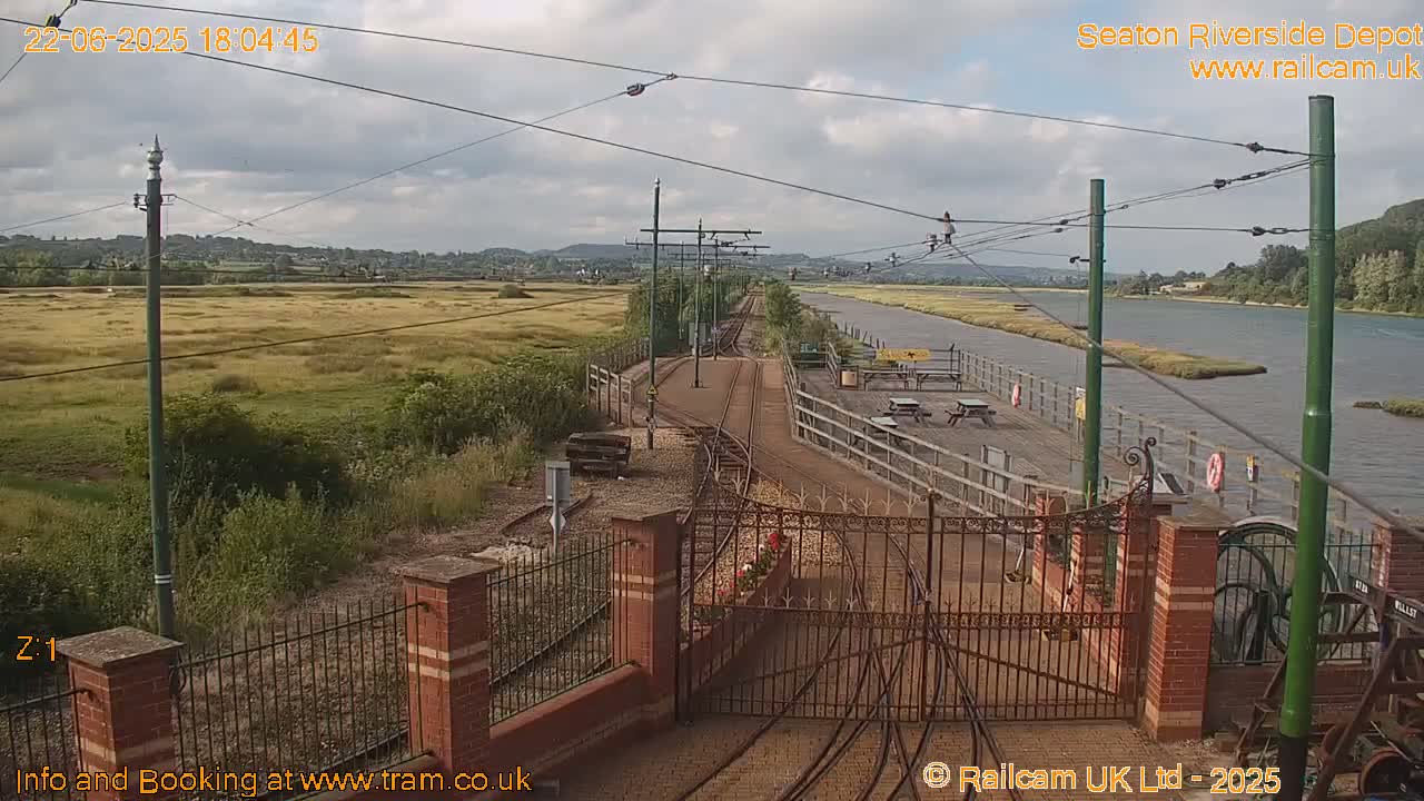 A train depot with tracks and a gate, situated beside a calm river under a partly cloudy sky.