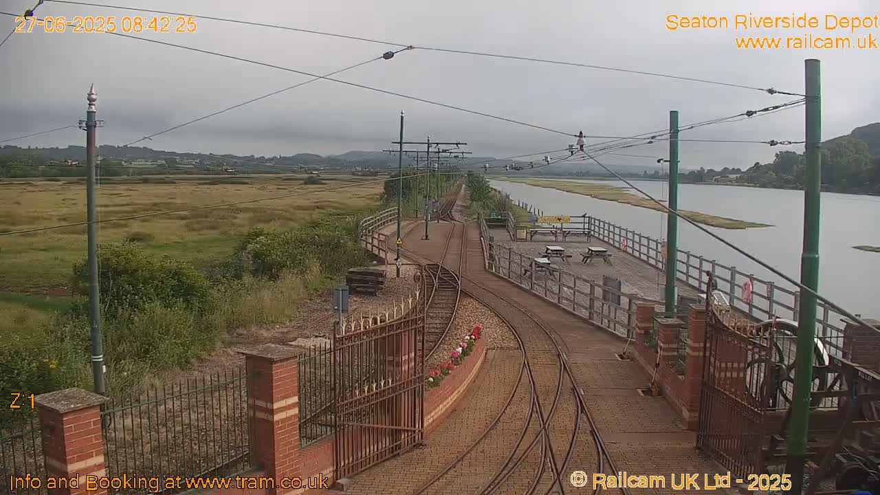 A railway track curves alongside a calm river under an overcast sky, leading to a small platform with picnic tables.