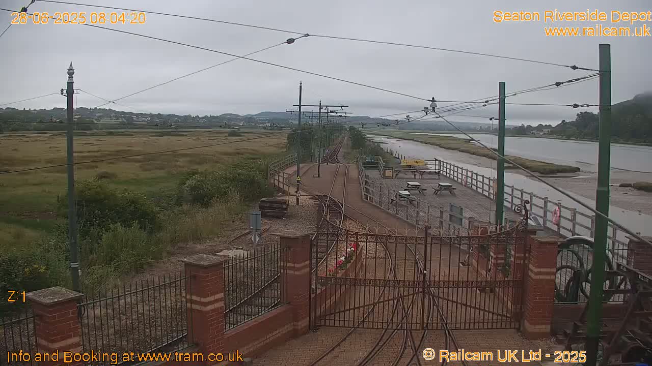A view of train tracks leading to a gated entrance, next to a body of water under an overcast sky.