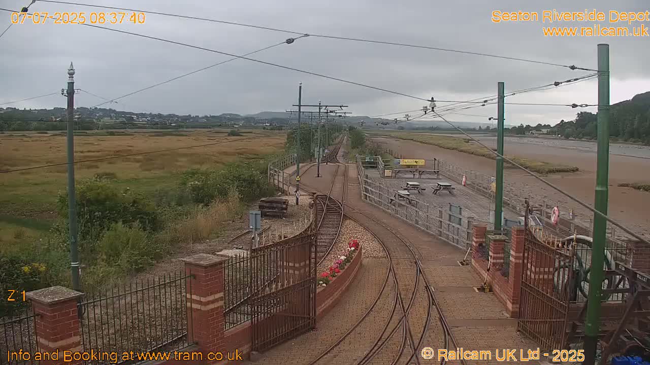 A railway depot with train tracks and a gated entrance, overlooking a wide expanse of flat land under an overcast sky.