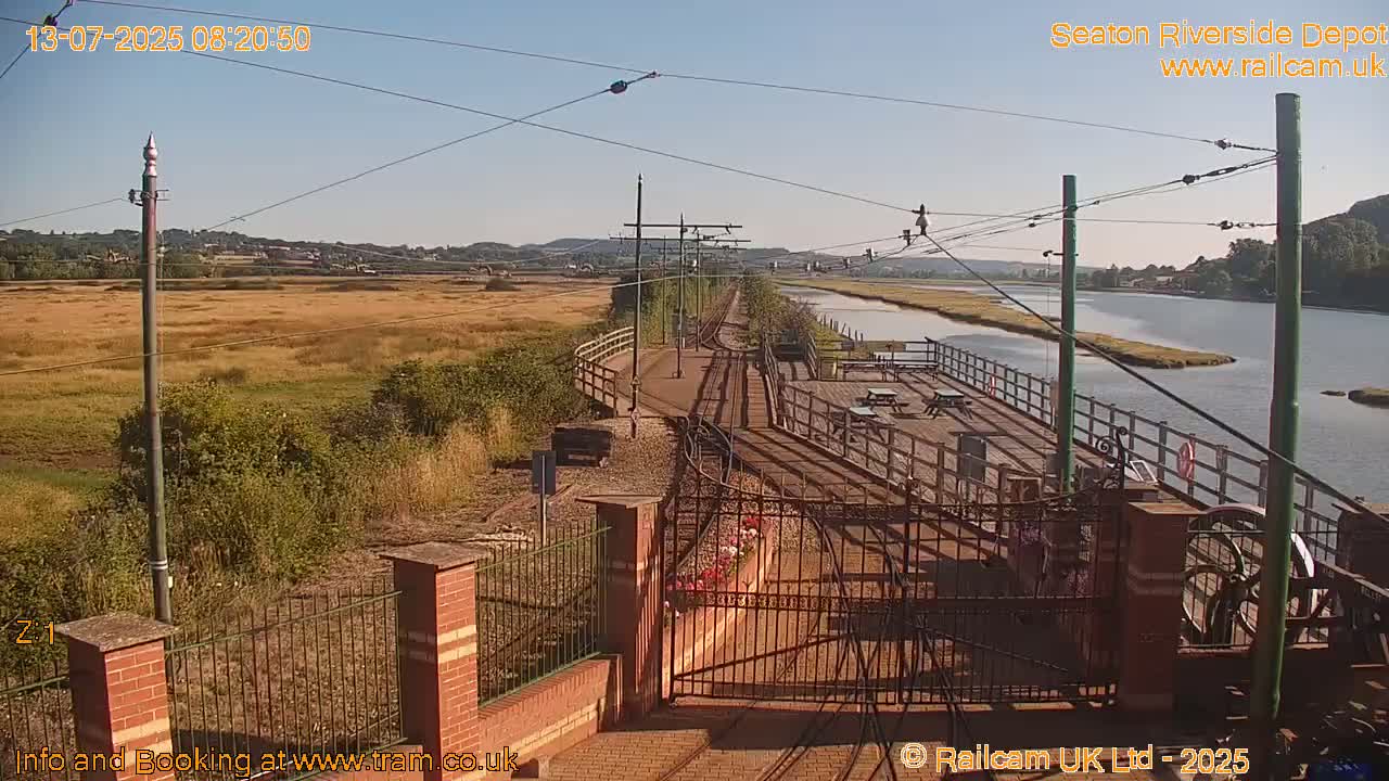 A sunny view of a train depot on a riverbank with a grassy field in the background.