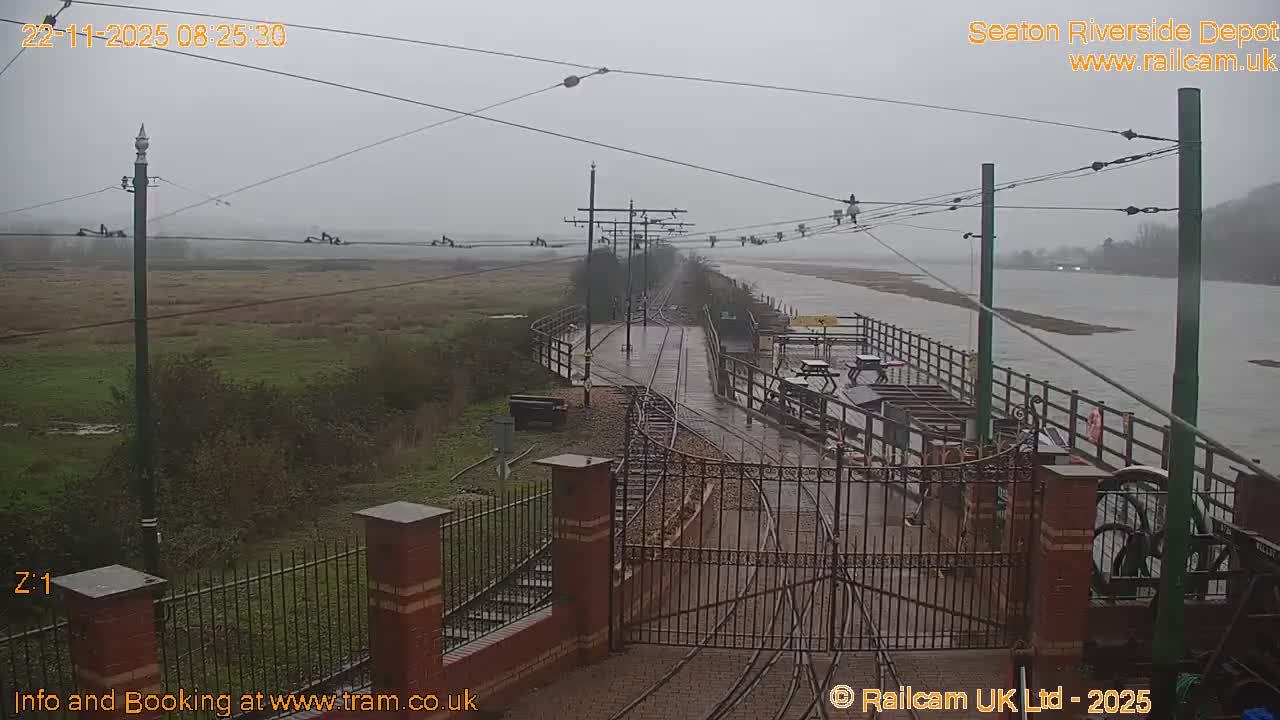 The image shows a wet tram line extending into a hazy distance alongside a wide river and expansive fields, all under an overcast, rainy sky.