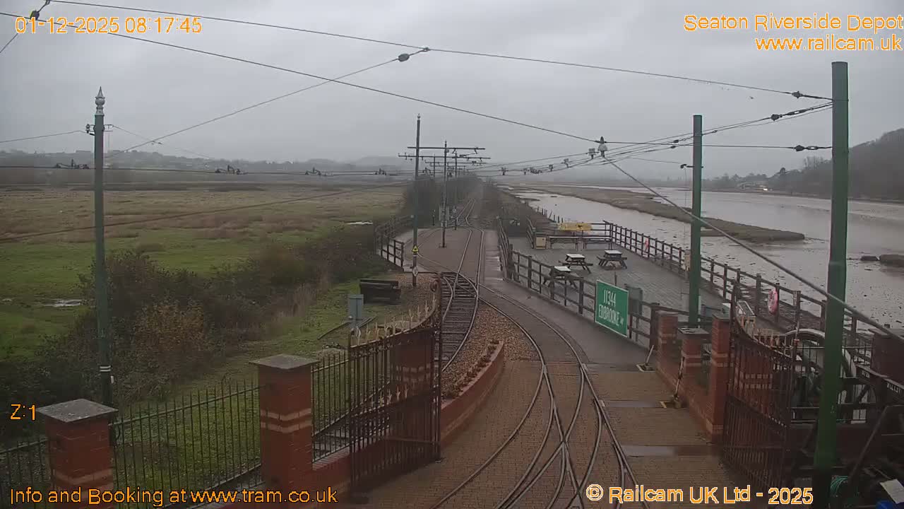 An elevated view shows a tramway with multiple tracks and a station platform featuring picnic tables, bordered by marshy fields and a wide, shallow river or estuary, all under a gray, overcast sky.