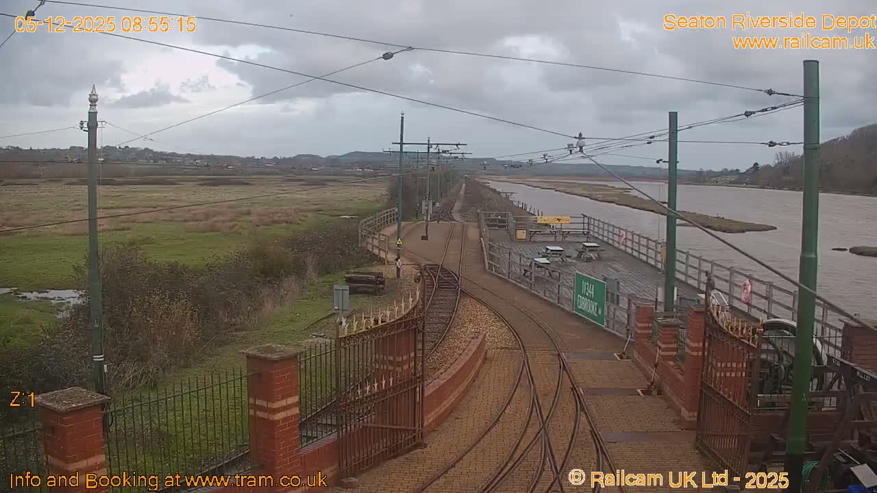 On a heavily overcast day, tram tracks curve in the foreground, flanked by a vast grassy marshland on one side and a wide river with a wooden pier and picnic tables on the other.