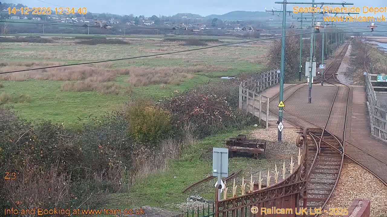 On a heavily overcast day, tram tracks curve in the foreground, flanked by a vast grassy marshland on one side and a wide river with a wooden pier and picnic tables on the other.