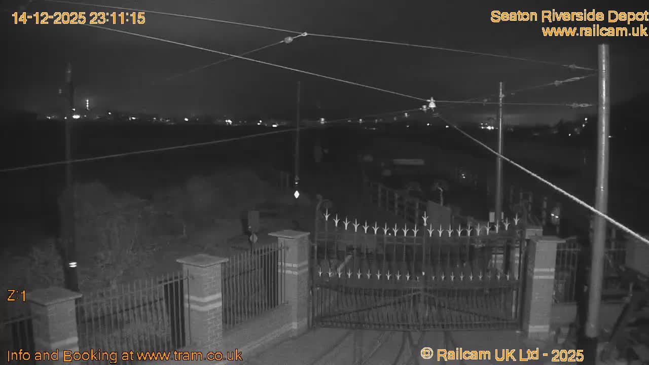 On a heavily overcast day, tram tracks curve in the foreground, flanked by a vast grassy marshland on one side and a wide river with a wooden pier and picnic tables on the other.