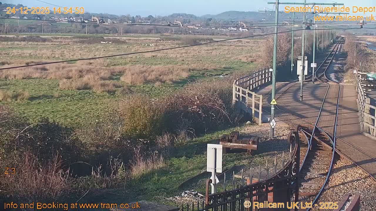 On a heavily overcast day, tram tracks curve in the foreground, flanked by a vast grassy marshland on one side and a wide river with a wooden pier and picnic tables on the other.