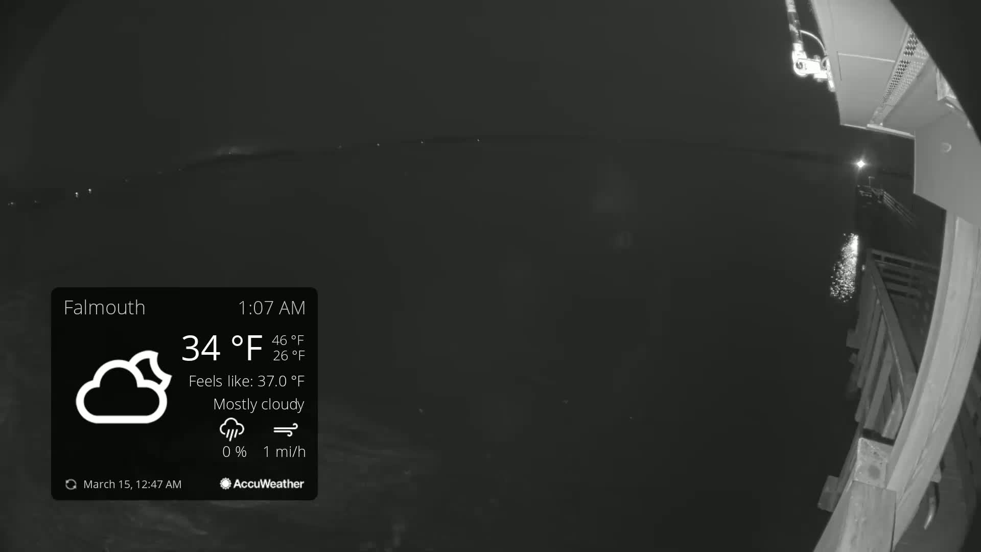 A nighttime, mostly cloudy scene shows a dark body of water with distant lights, viewed from a wooden deck under a 34°F temperature.