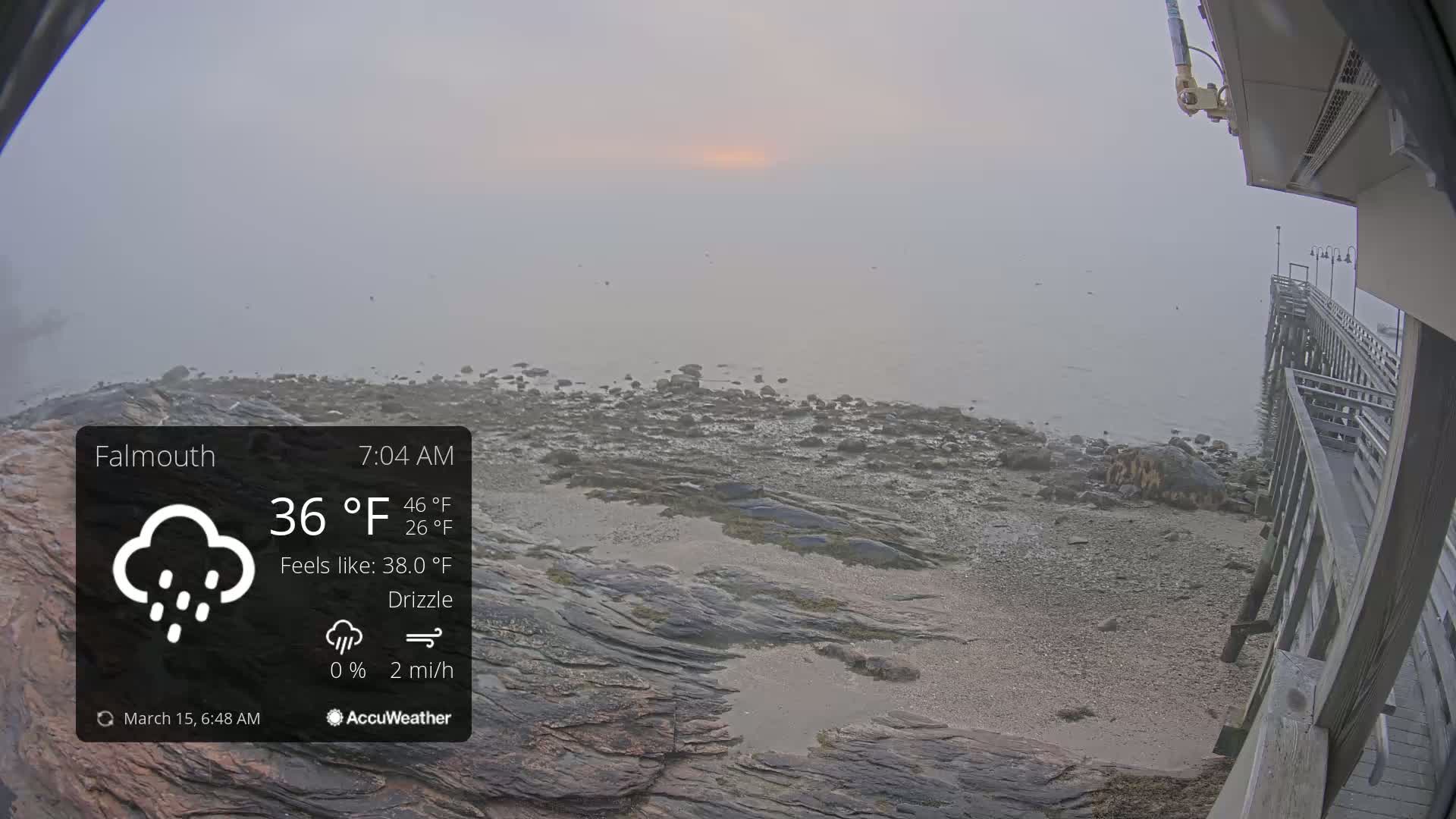 A foggy, drizzly morning scene shows a rocky shoreline with a wooden pier extending into calm water.