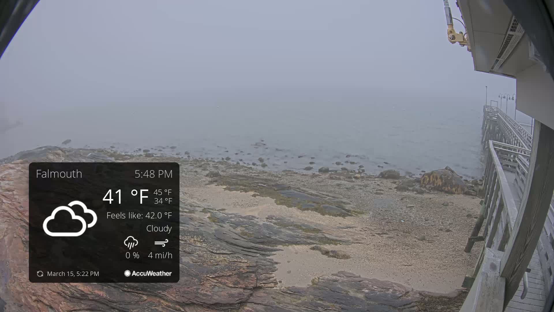A foggy, cloudy view of a rocky shoreline and a wooden pier extends into a calm body of water.