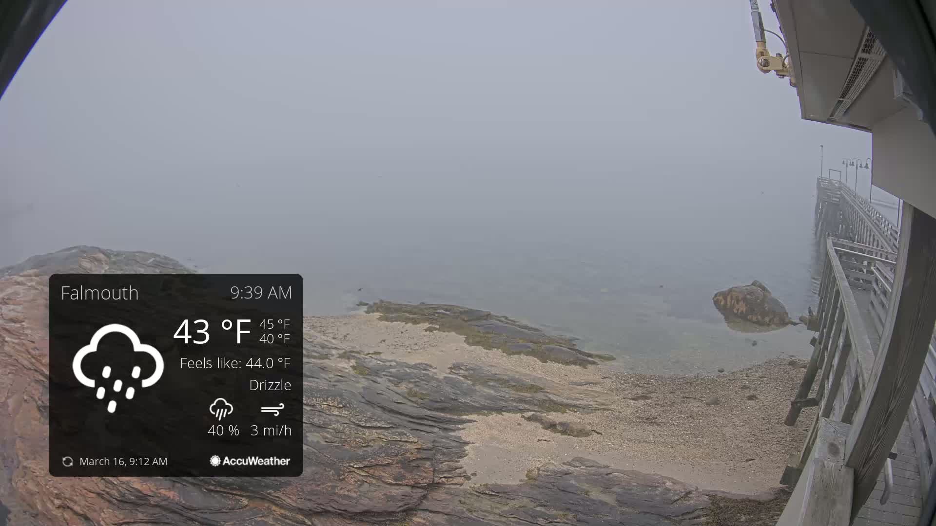 A foggy, drizzly scene shows a rocky shoreline and a pier extending into the water.