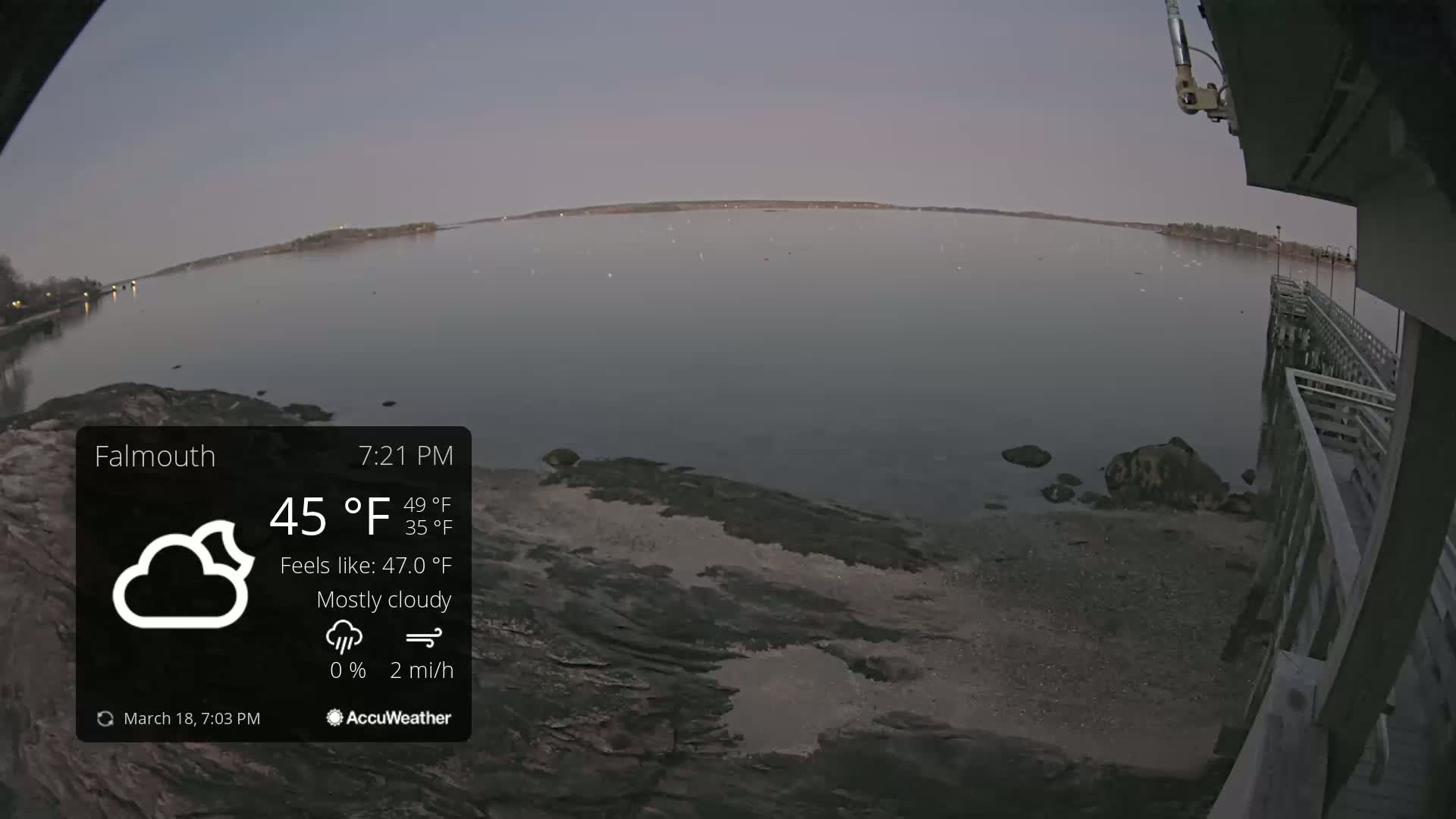 A mostly cloudy evening scene shows a calm body of water with a rocky shoreline and a wooden pier under a twilight sky.