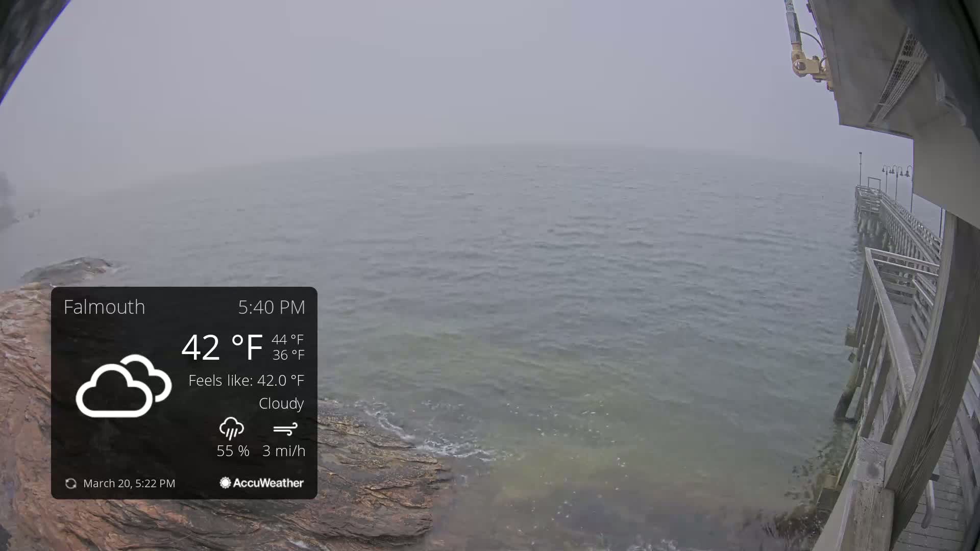 A cloudy, 42°F day shows a view of a body of water from a wooden pier with light waves lapping at the rocky shore.