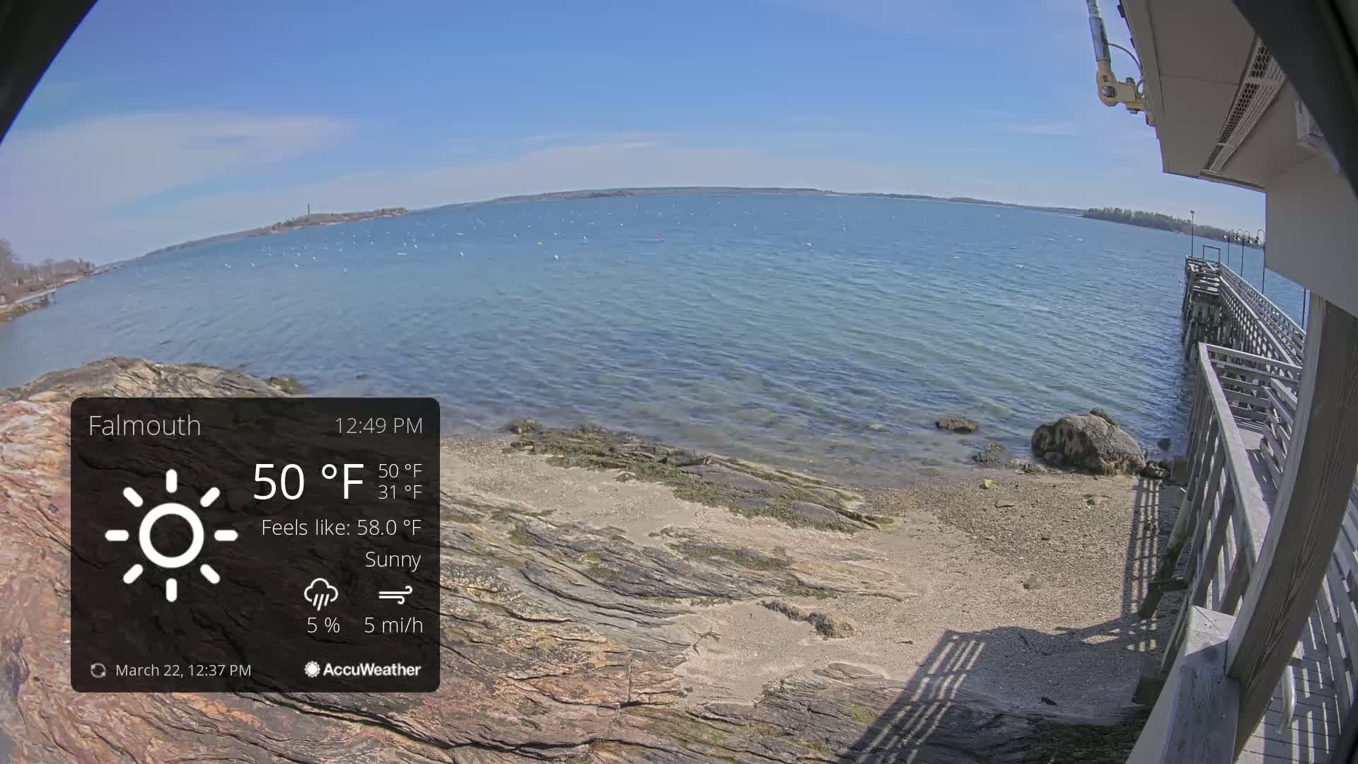A sunny, partly cloudy day at a rocky shoreline with a wooden pier extending into calm, blue water.