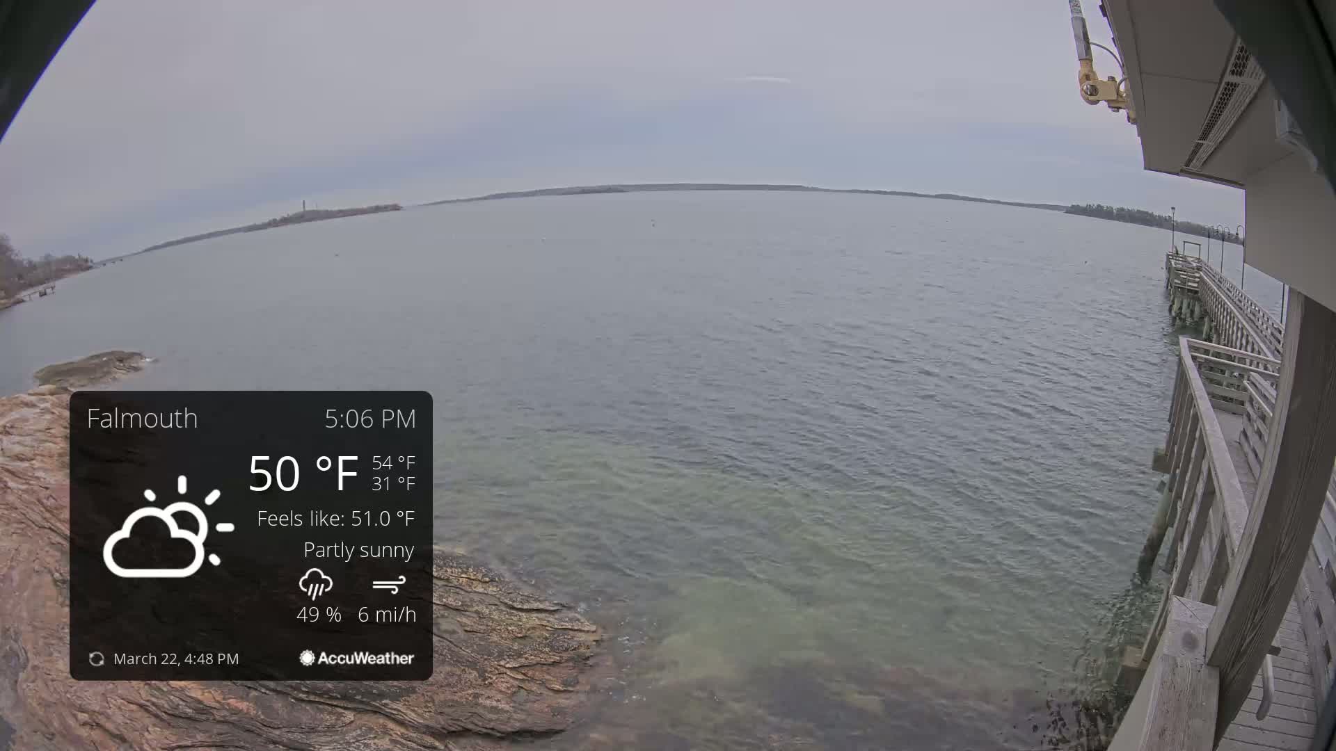 A partly sunny view of a calm body of water from a wooden pier, with a rocky shoreline visible in the foreground.